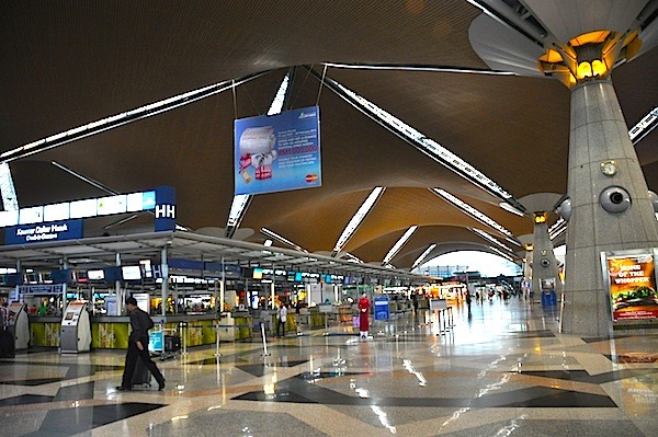 Interior view of Kuala Lumpur International Airport (KLIA) terminal with modern architecture, check-in counters, passengers walking, and spacious seating area.